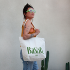 Person holding a white tote bag with green Basalt Logo, standing against a plain background with a cactus plant.